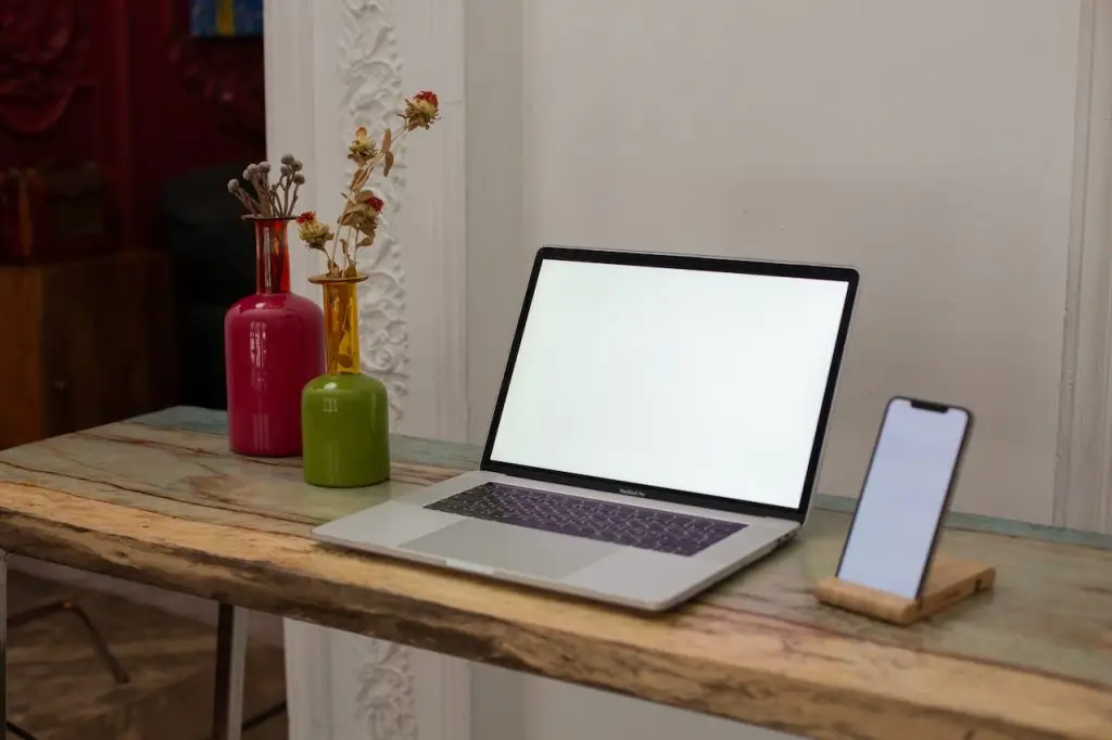 A MacBook with a blank screen and an iPhone on a wooden stand are placed on a rustic wooden table. Decorated vases with dried flowers sit to the left of the laptop. The background features a white ornate wall.
