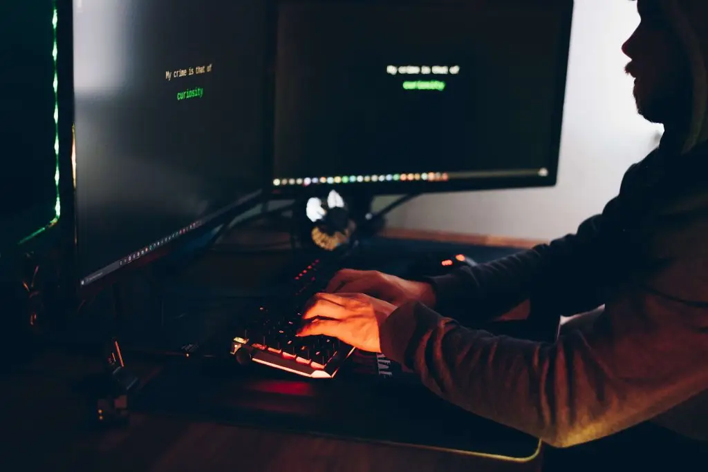 A person in a dark hoodie is typing on a keyboard at a dimly lit desk with two large monitors displaying green text. The surrounding area has a low light ambiance, emphasizing the screens and the typing hands.