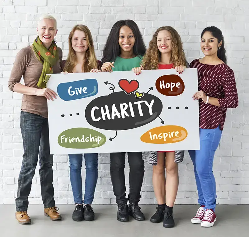 A diverse group of five people stands together against a white brick wall, smiling and holding a large sign reading "CHARITY" with icons around it. The icons say "Give," "Friendship," "Hope," and "Inspire." The mood is positive and encouraging, reflecting the camaraderie often seen in managed services teams.