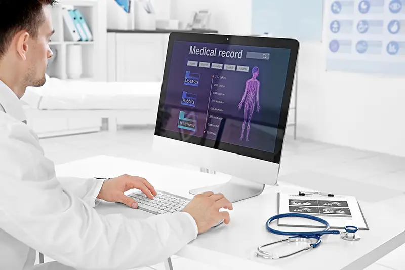 A medical professional sits at a desk, using a computer with Managed IT Services. The screen displays a digital medical record, including a human body diagram and various health metrics. Nearby are a stethoscope, clipboard with documents, and medical books. The setting is an examination room.