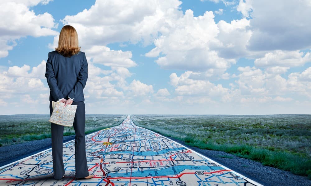 The image displays a businesswoman standing on a road that looks like a map and looking forward to the next step.