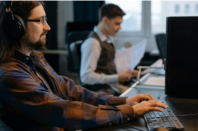 Two professionals in an office setting providing remote IT support. One focused on a computer screen with headphones, and the other reviewing documents in the background, exemplifying teamwork in tech support services.