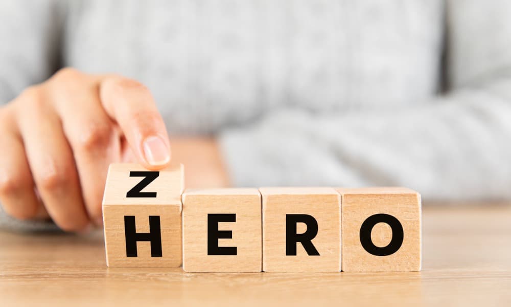 A woman’s hand is on wooden tiles with letters, tipping one so that the word zero turns into the word hero.
