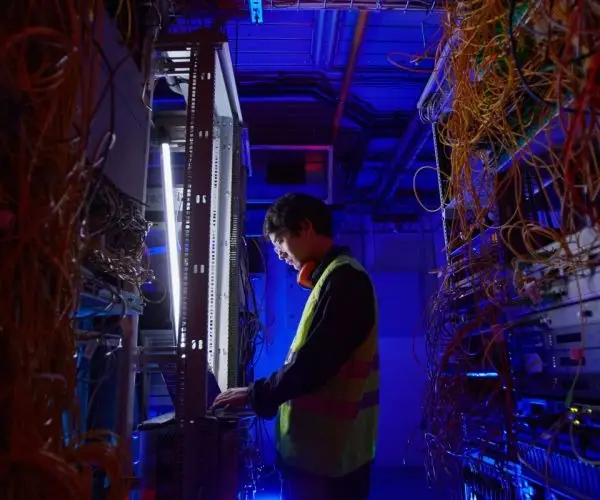 A technician in a high-visibility vest works on a server rack in a dimly lit data center in Seattle, surrounded by numerous cables and equipment. The scene, bathed in blue and purple lighting, highlights the complexity of the technological environment typical of IT consulting projects.