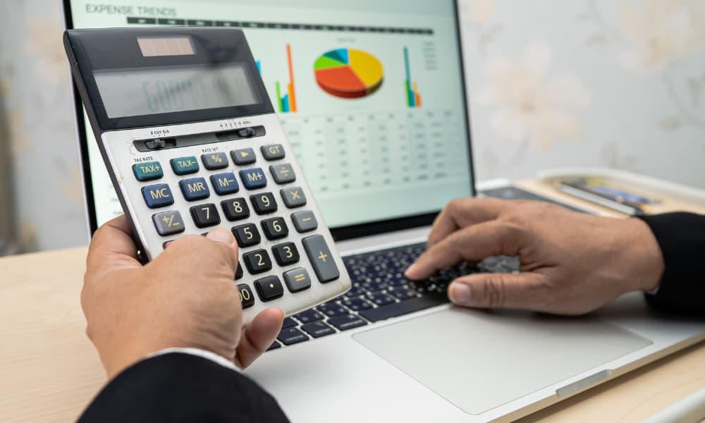A person in a suit deftly navigates both a calculator and a laptop, the screen alive with colorful charts labeled "EXPENSE TRENDS." In this office setting, they analyze I.T. Budget data to avoid wasting money, ensuring every decision supports growth.