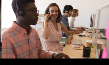 A diverse group of customer service representatives working at computers, wearing headsets. A woman in the center smiles, creating a friendly atmosphere.