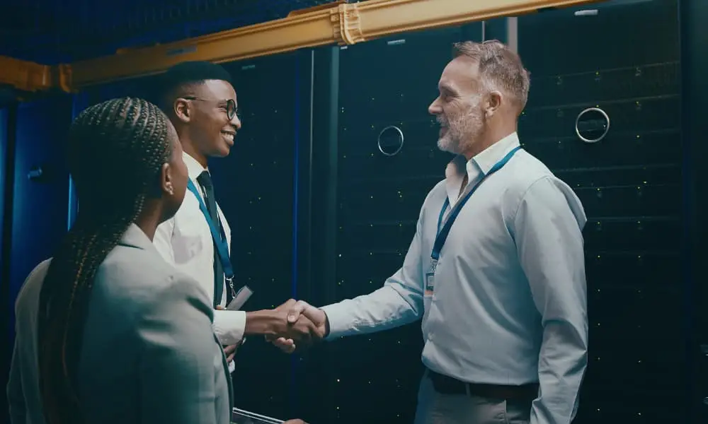 Three professionals meeting beside server racks, with two shaking hands.