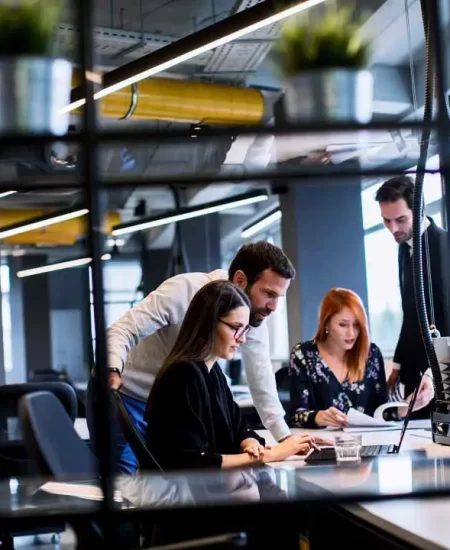 A group of four people in a modern office setting collaborate around a laptop. Two are standing and two are seated, engaged in a discussion. The room features industrial design elements and large windows letting in natural light.